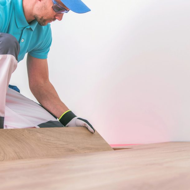 A worker in a blue shirt and protective gloves is installing a wooden floor, bending over to carefully place or adjust a plank.